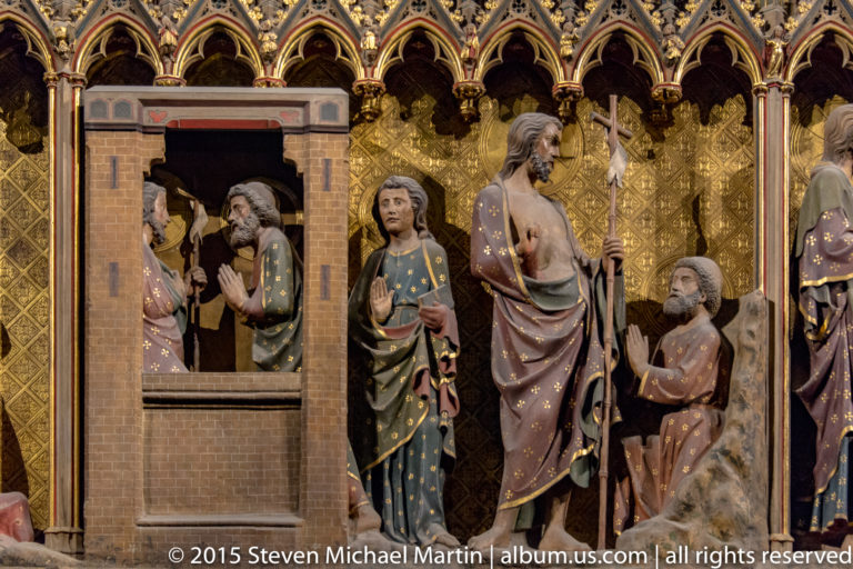 Christ stands beside a small walled building as nearby painted figures kneel and gesture toward him.