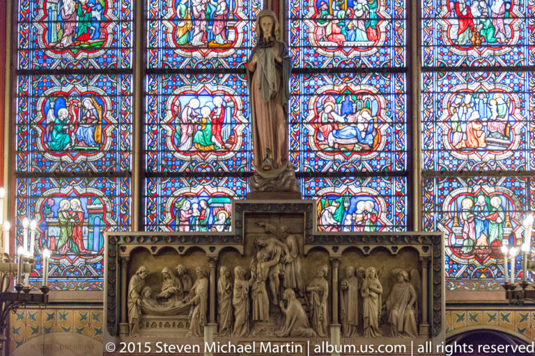 Standing religious sculpture centered before multicolored stained glass, with carved relief panels and stone figures below.