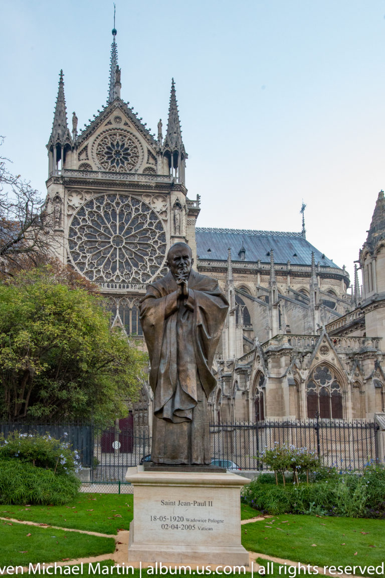 Bronze statue of Saint John Paul II in a garden with Notre-Dame’s rose window and south transept behind.