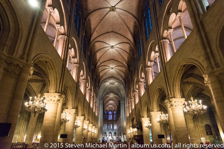 Centered nave view with rib vaults, pointed arcades, chandeliers, columns, and distant stained glass.