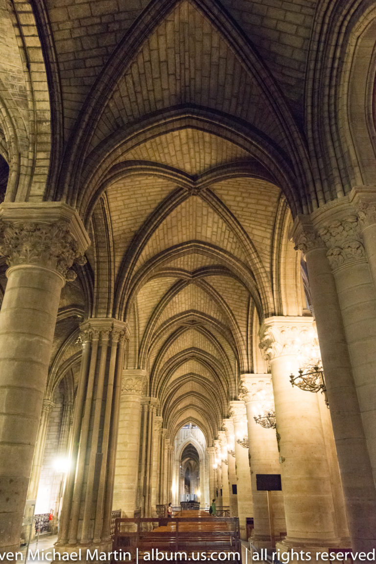 Long stone side aisle with repeated rib vaults, heavy piers, arches, and wall-mounted chandeliers.