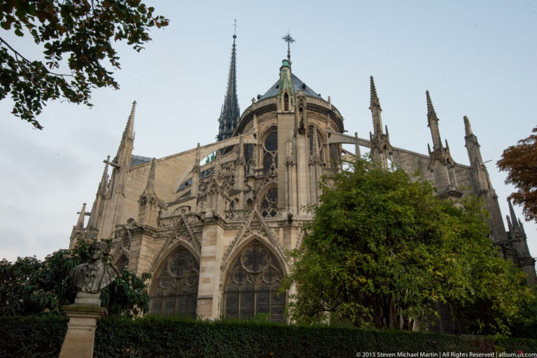 Garden view of Notre-Dame’s apse with flying buttresses, pinnacles, radiating chapels, spire, and trees.