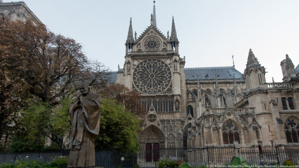 Garden statue in front of Notre-Dame’s south transept, with rose window, gable, pinnacles, and buttresses behind.