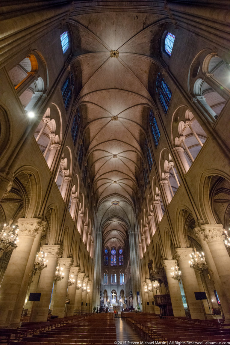 Wide interior view down the nave with chairs, columns, arches, chandeliers, clerestory windows, and rib vaults.