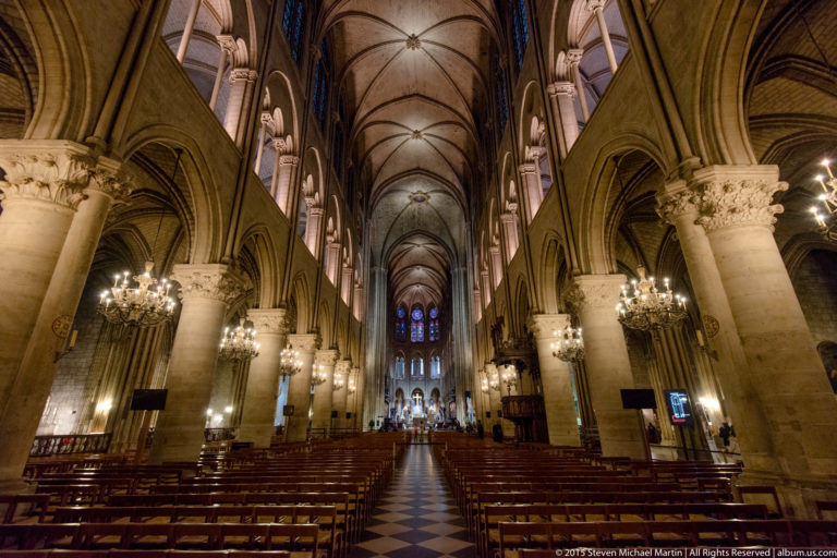 Centered nave view with rows of chairs, columns, pointed arcades, chandeliers, rib vaults, and distant choir.