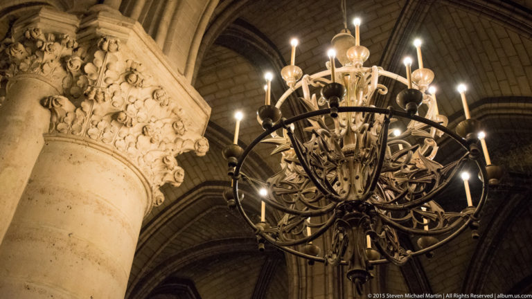 Large circular metal chandelier with many candle-like lights beside a carved foliated stone capital.