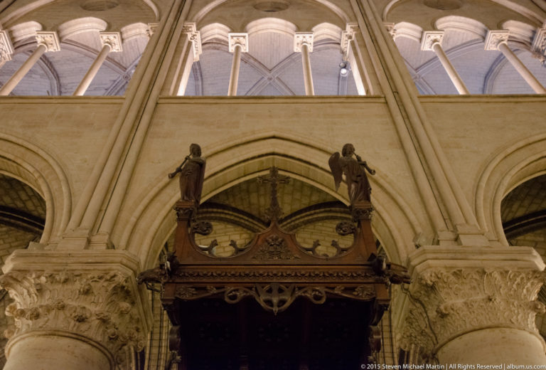 Dark carved wooden canopy between large stone piers with foliated capitals and pointed gallery arches above.