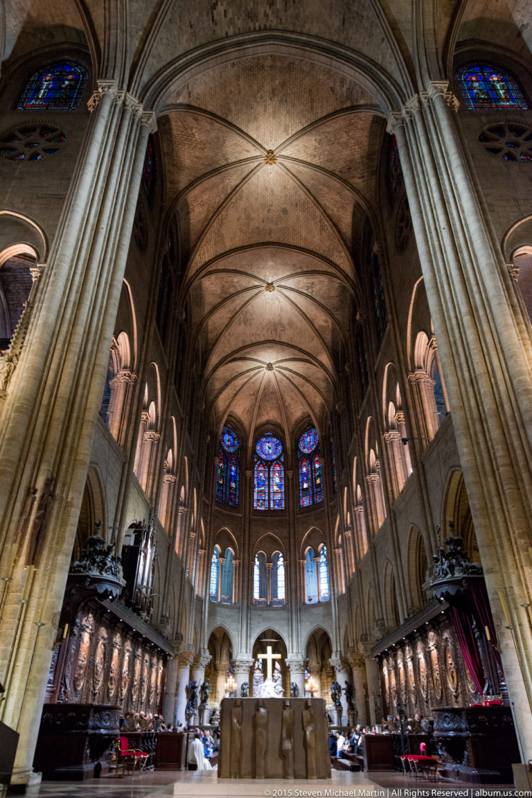Centered choir view with altar and cross, apse windows, sculpted choir screens, columns, and high vaults.