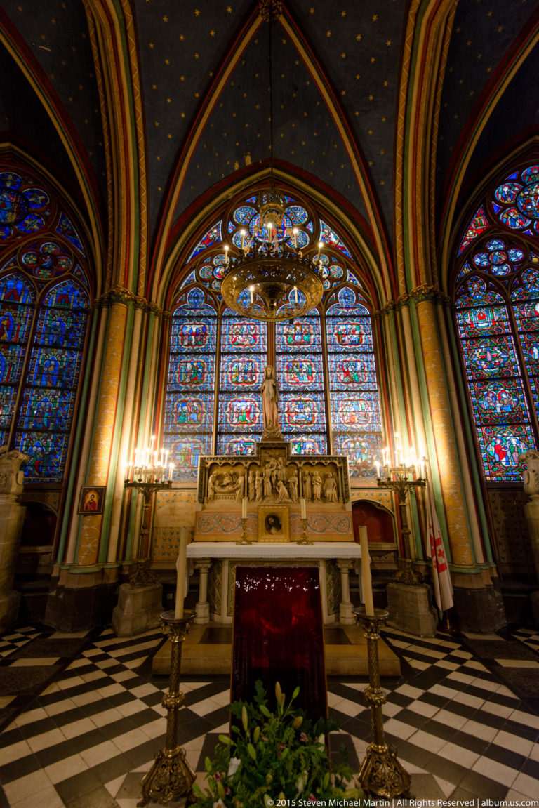 Wide chapel view with central altar, stained glass, star-painted vault, checkered floor, candles, and chandelier.