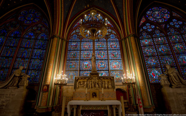 Side chapel with a sculpted altar, chandelier, tall stained-glass windows, candles, and flanking statues.