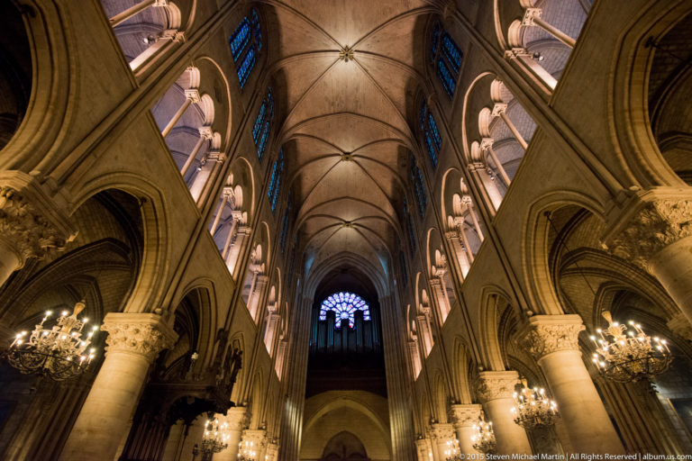 Long nave view toward the west rose window and organ, framed by columns, arcades, chandeliers, and rib vaults.