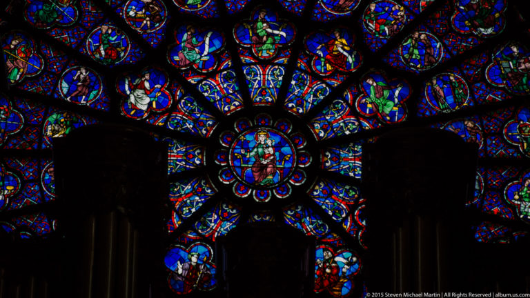 Close view of a rose window with radiating colored glass panels and a central figural medallion.