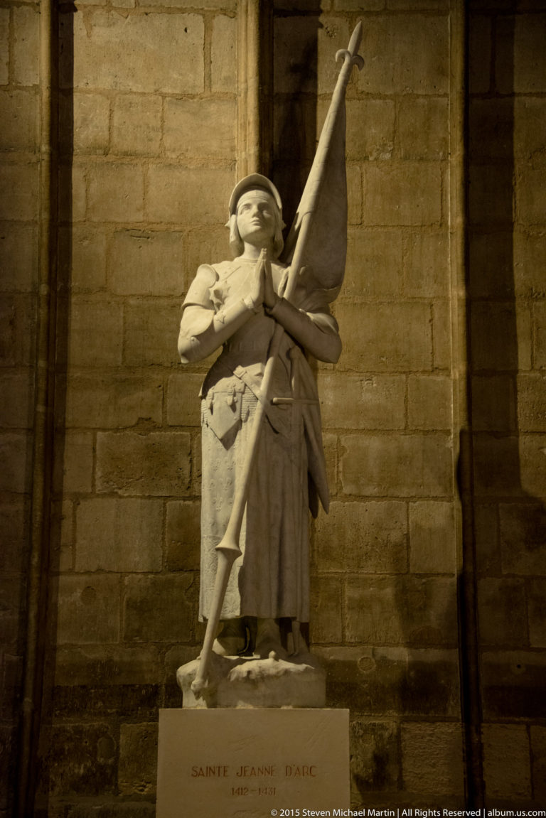 Marble statue of Saint Joan of Arc in armor on an inscribed pedestal inside Notre-Dame de Paris.