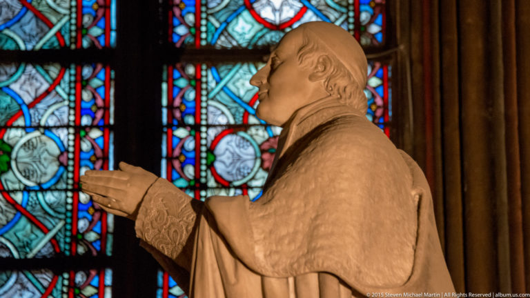 Profile view of a pale clerical statue with hands raised before multicolored stained-glass windows.