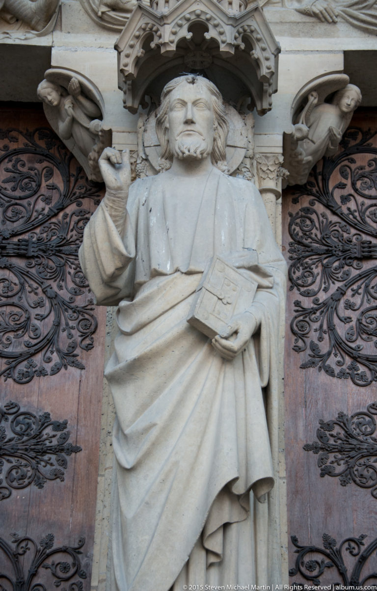 Stone robed figure beside a carved wooden portal door, one hand raised and the other holding an open book.