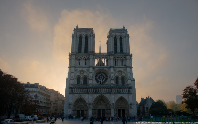 Frontal dusk view of Notre-Dame’s west façade with twin towers, rose window, three portals, and people below.