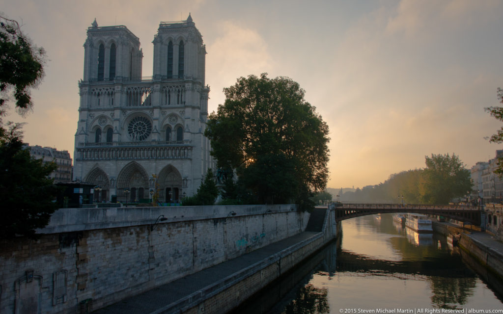 Shadowed west façade of Notre-Dame beside the Seine at sunset, with twin towers, rose window, and river.
