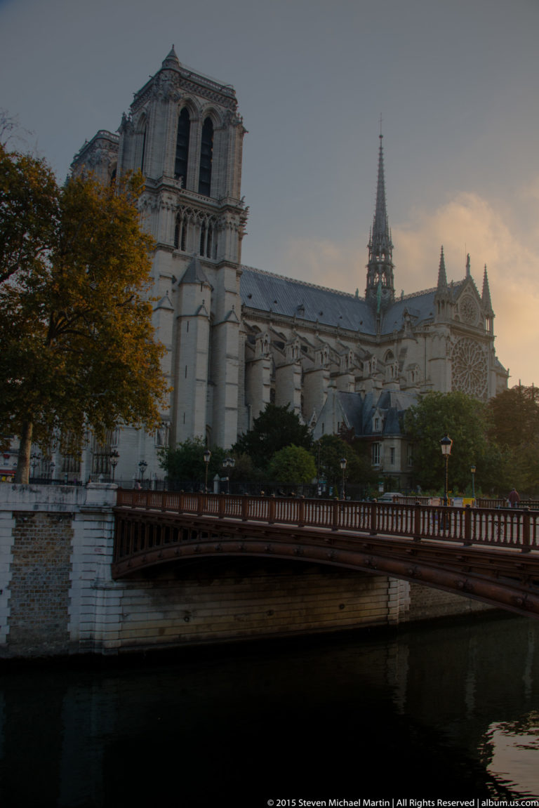 Notre-Dame beyond a bridge over the Seine, with towers, spire, trees, and embankment walls.