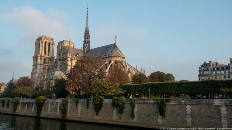 Notre-Dame across the Seine with twin towers, spire, apse, autumn trees, and long stone embankment wall.