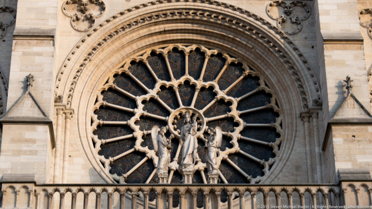 Close exterior view of Notre-Dame’s west rose window with radiating tracery and central sculpted figure.