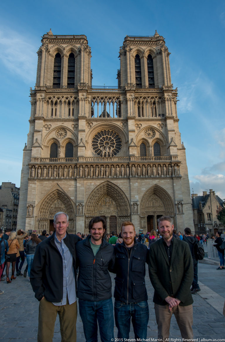 Four men standing together in front of Notre-Dame’s west façade with twin towers and rose window.