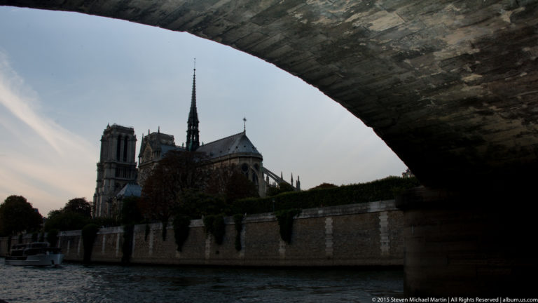 Notre-Dame viewed from the Seine beneath a dark bridge arch, with towers and spire beyond the river wall.