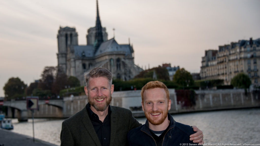 Two men stand arm in arm near the Seine with Notre-Dame softly focused behind them.