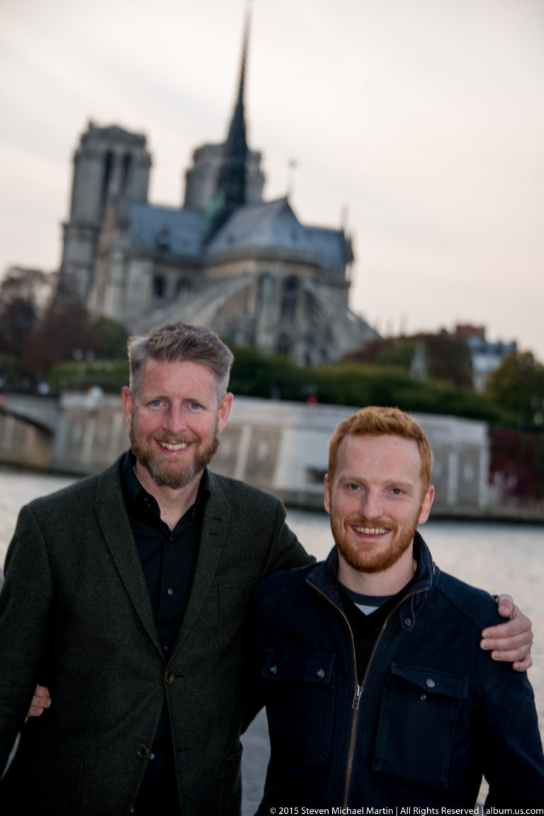 Two smiling men pose beside the Seine with Notre-Dame de Paris blurred in the background.