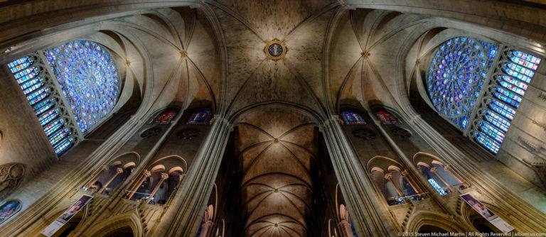 Upward interior view of clustered piers, rib vaults, and large rose windows glowing on both sides.