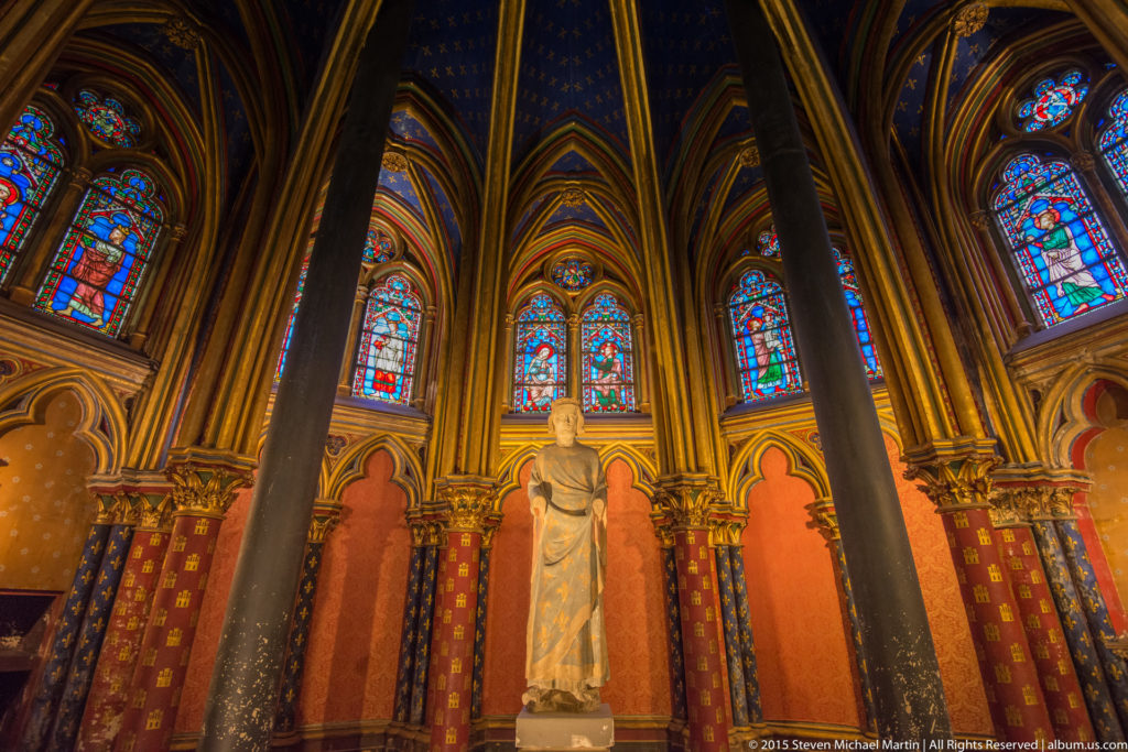 Lower chapel of Sainte-Chapelle with a pale stone statue between dark columns, beneath stained-glass windows and blue-and-gold vaulting.