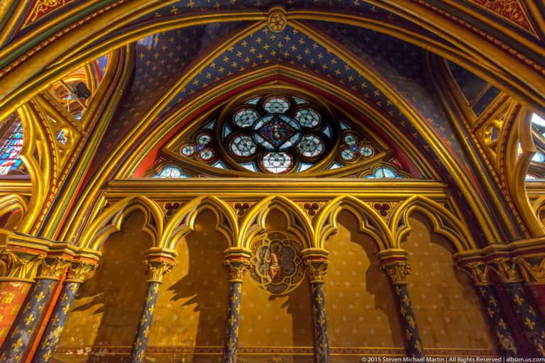 Lower-chapel wall detail at Sainte-Chapelle with a quatrefoil window, blind pointed arcade, heraldic medallion, and painted gold-star decoration.