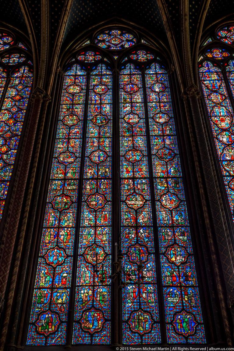 Towering stained-glass lancet windows in Sainte-Chapelle’s upper chapel, divided by narrow dark masonry shafts beneath traceried arches.