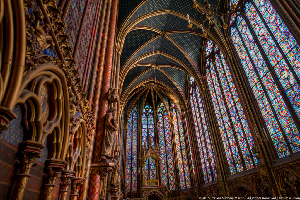 Oblique view through Sainte-Chapelle’s upper chapel toward the apse, with stained-glass walls, slender supports, carved arcades, and ribbed vaults.