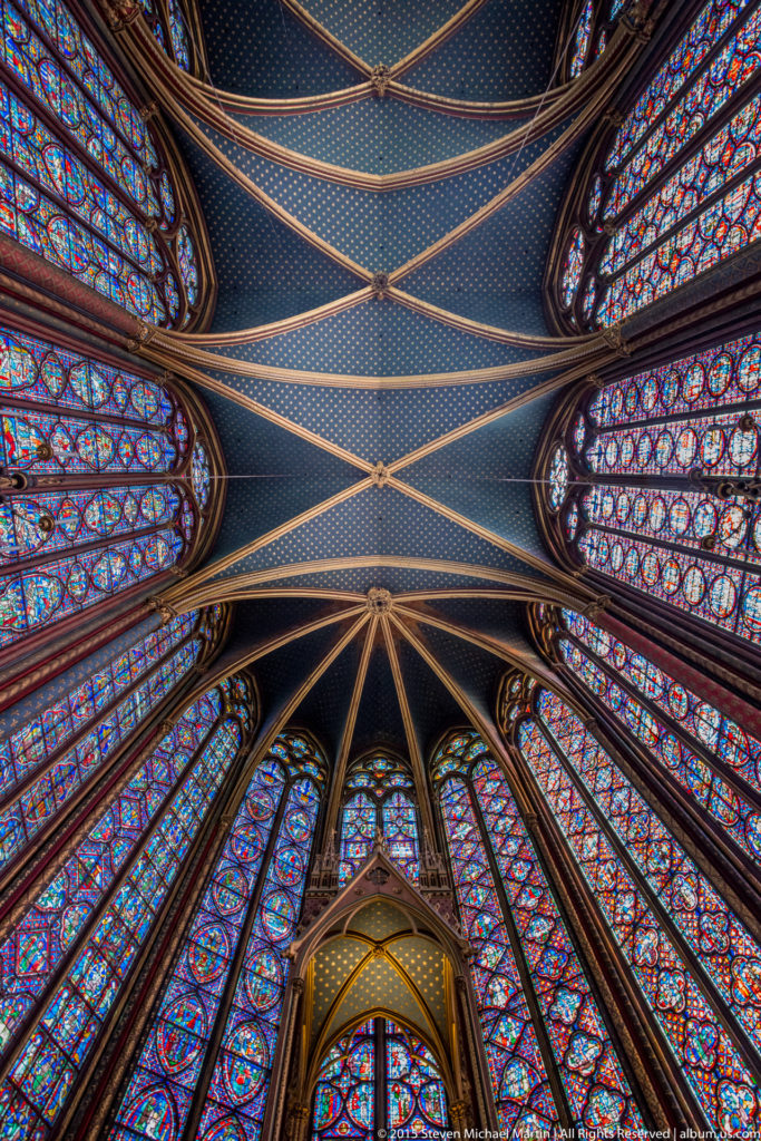 Symmetrical upward view of Sainte-Chapelle’s upper-chapel vault, framed on both sides by stained-glass windows.