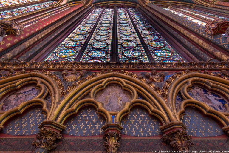 Upper-chapel wall at Sainte-Chapelle with stained-glass lancets above a carved blind arcade containing painted quatrefoil panels.