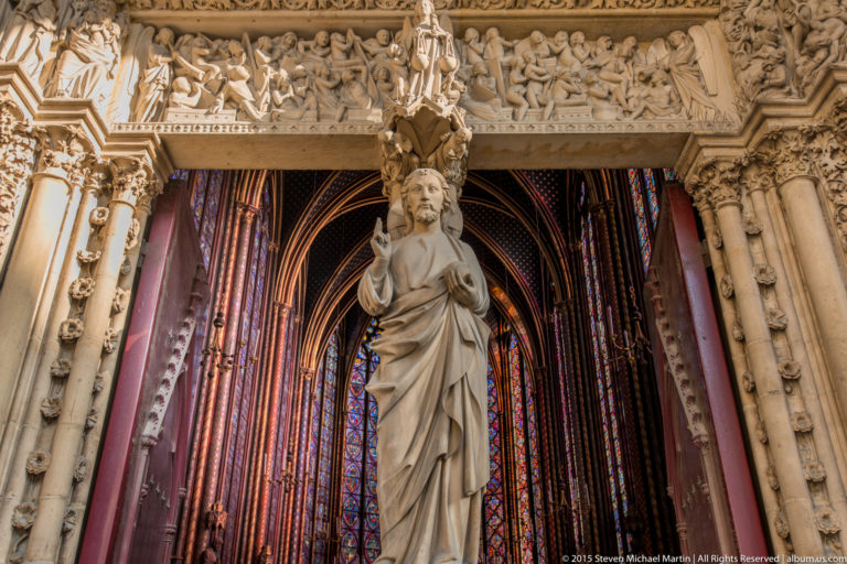 Stone apostle statue in Sainte-Chapelle’s upper chapel, standing before stained-glass windows and richly carved architectural ornament.