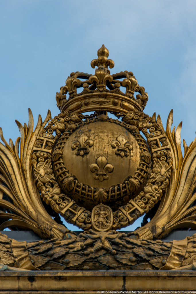 Gilded royal emblem with crown, fleurs-de-lis, and the collar of the Order of Saint Michael on an exterior architectural crest near Sainte-Chapelle.