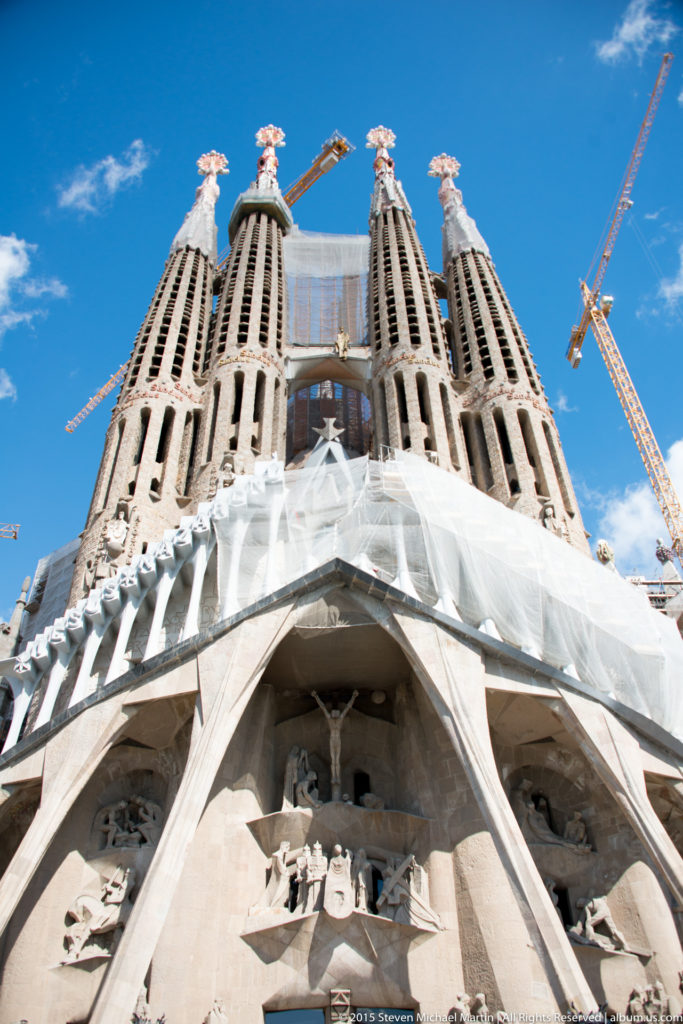 Low-angle view of the Sagrada Família Passion façade with inclined portico columns, four bell towers, protective sheeting, and cranes.