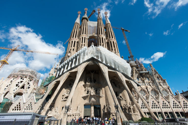 Street-level view of the Sagrada Família Passion façade with inclined columns, angular sculpture, four towers, visitors, and cranes.