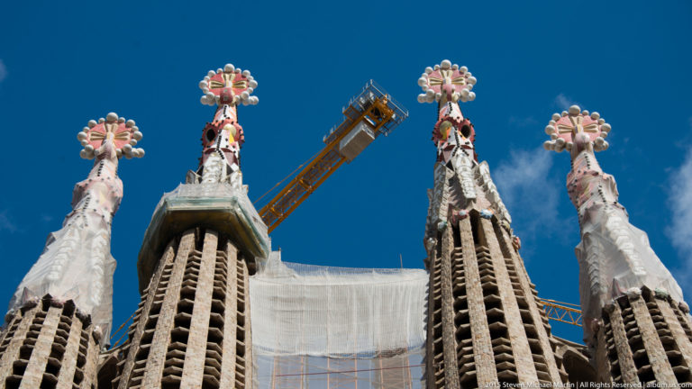 Low-angle view of colorful floral and cross-shaped pinnacles atop Sagrada Família towers, with a construction crane between them.