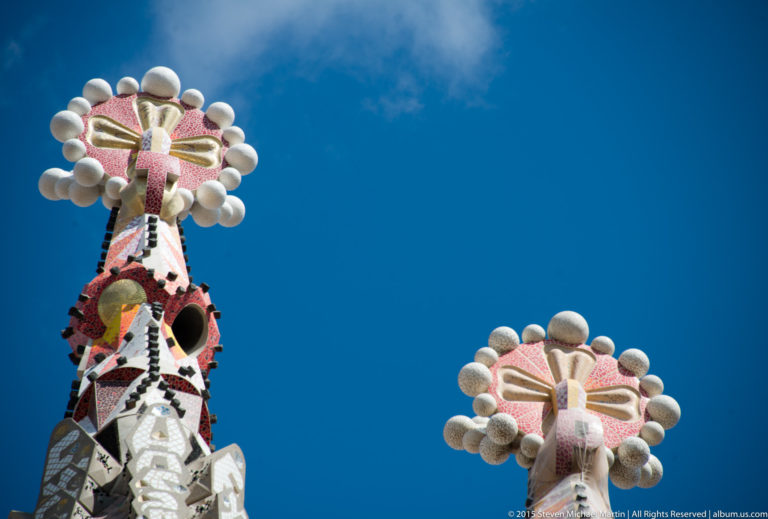Two colorful mosaic-covered Sagrada Família tower pinnacles with rounded white ornaments against a blue sky.