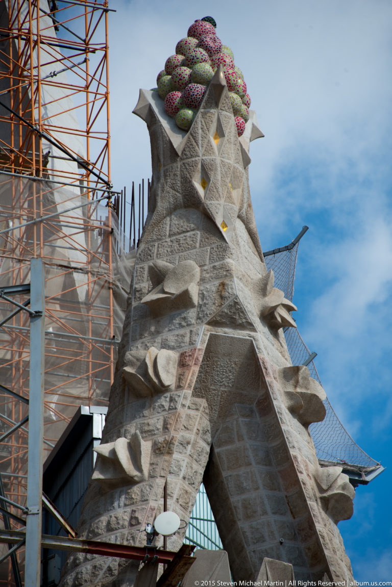 Faceted stone pinnacle crowned with colorful fruit-like mosaic forms at the Sagrada Família, with scaffolding alongside.