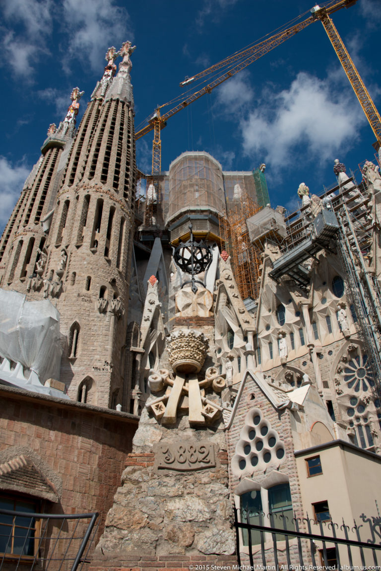 Oblique Sagrada Família exterior with tower, sculptural stonework, circular openings, scaffolding, platforms, and cranes.