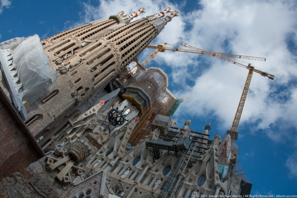 Steep upward view of a Sagrada Família tower beside cranes, scaffolding, temporary structures, and sculptural stonework.