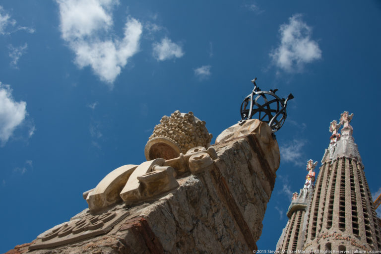 Low-angle Sagrada Família detail with projecting stone ornament, dark openwork metal form, and a decorated tower behind.