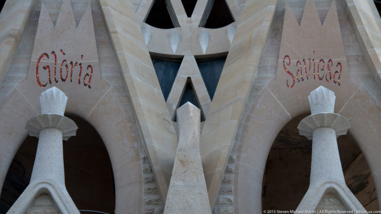 Red inscriptions reading “Glòria” and “Saviesa” on pale geometric stonework with columns and angular openings.