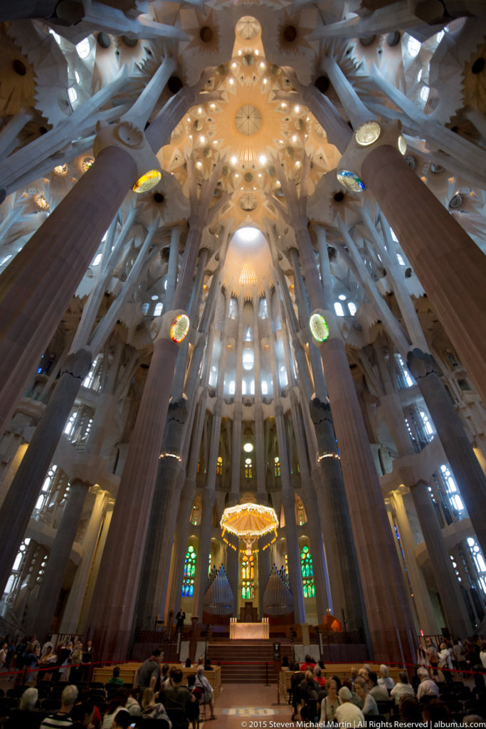 Symmetrical Sagrada Família nave with tall branching columns framing the sanctuary beneath a bright patterned vault.