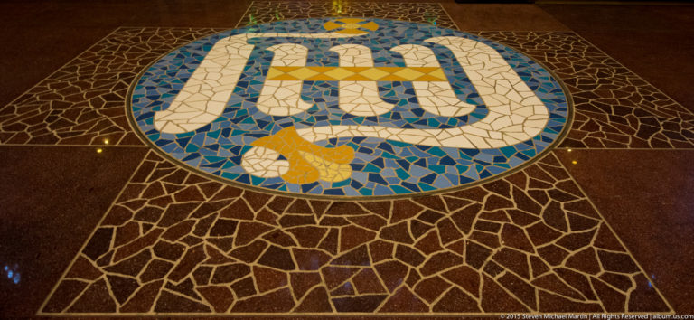 Circular blue, white, and yellow floor mosaic bearing the letters JHS within irregular dark stone paving.