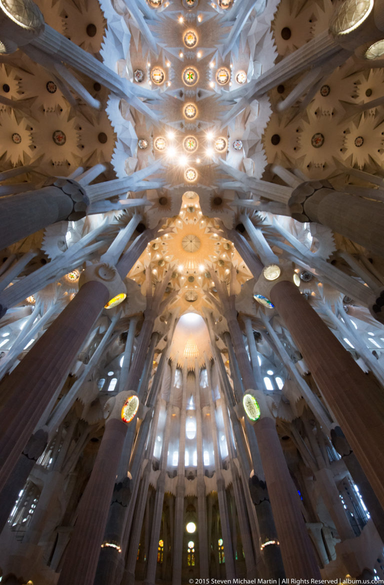 Upward Sagrada Família nave view with branching supports, geometric vault panels, and a central line of glowing circular ceiling elements.