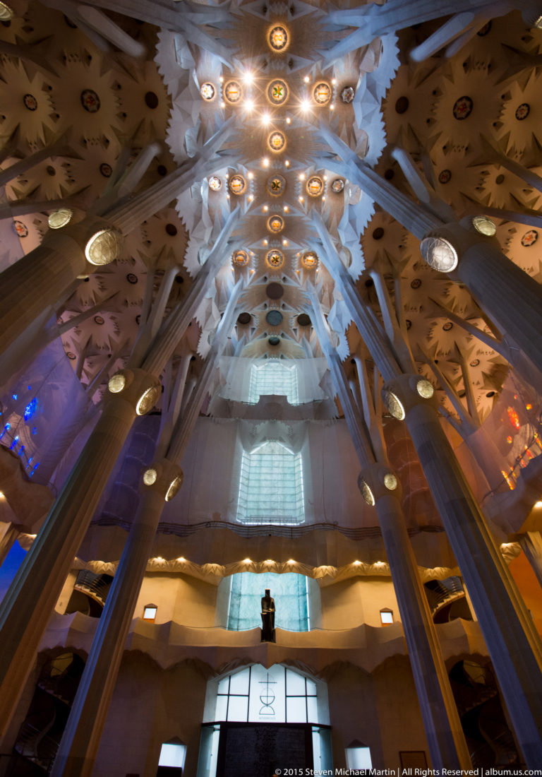 Low-angle Sagrada Família interior with branching columns, galleries, bright entrance glazing, and illuminated ceiling forms.